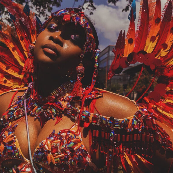 The author poses for a phoo while wearing a costume inspired by the Chinese dragon during Trinidad Carnival. The costume features many feathers, gems and tassels of varied hues of red, orange, yellow, orange, and touches of blue.