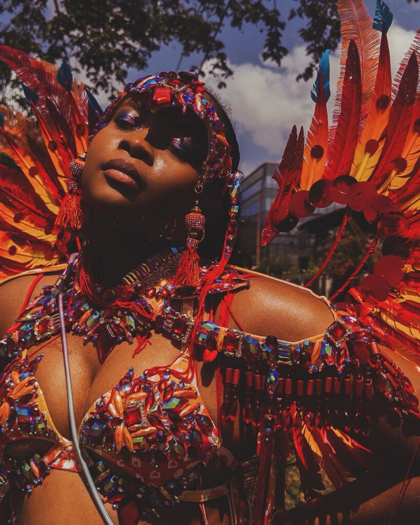 The author poses for a phoo while wearing a costume inspired by the Chinese dragon. The costume features many feathers, gems and tassels of varied hues of red, orange, yellow, orange, and touches of blue.