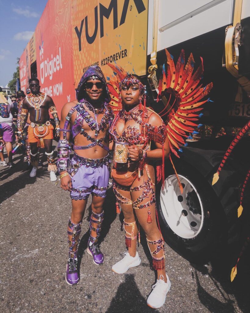 The author poses for a photo with a friend while wearing a costume inspired by the Chinese dragon. The costume features many feathers, gems and tassels of varied hues of red, orange, yellow, orange, and touches of blue.