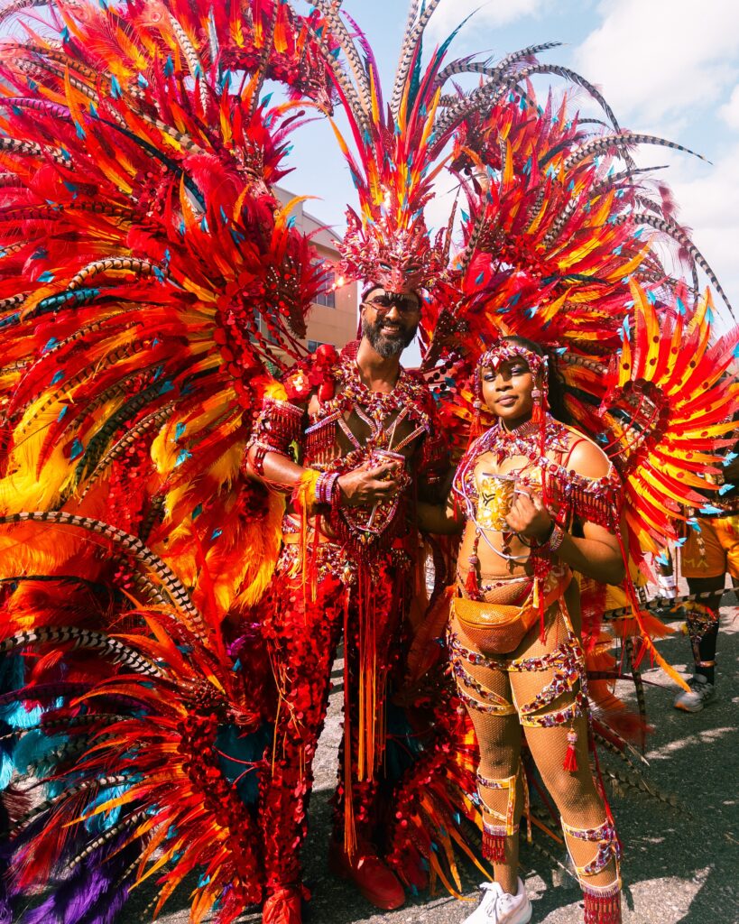 The author poses for a photo with Rawle Permanad, the designer of her Carnival costume, while wearing a costume inspired by the Chinese dragon. The costume features many feathers, gems, and tassels in varied hues of red, orange, yellow, and blue.